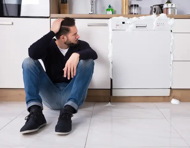 Man sitting on the ground beside his broken dishwasher, frustrated.