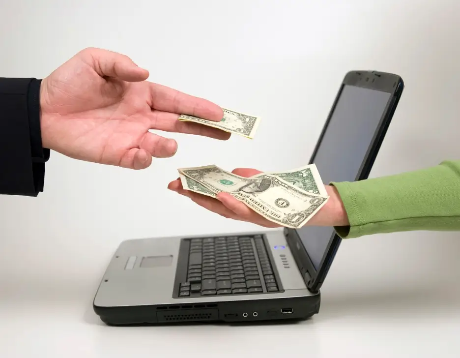Two hands exchanging money in front of a laptop computer.