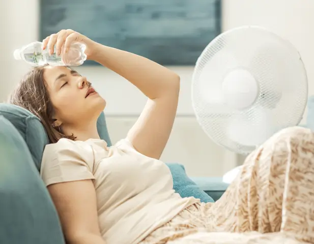 A woman sitting on her couch, holding an ice pack on her forward, with the floor fan running beside her.