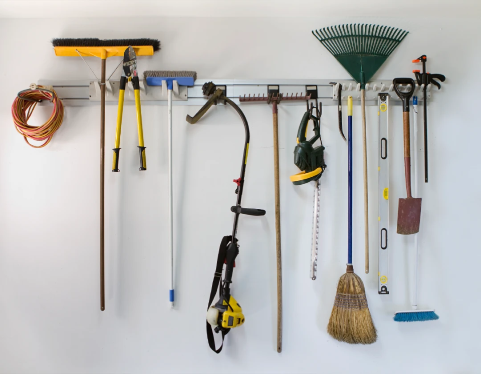 Organized garage wall-mounted tool rack holding garden tools including a rake, brooms, string trimmer, loppers, shovel, level, and extension cord