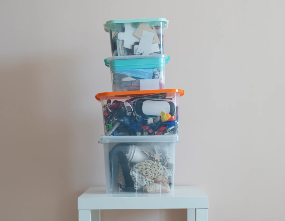 Three clear plastic storage bins with colorful lids stacked on a white side table, filled with craft supplies, accessories, and household items