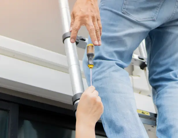 A homeowner standing on a ladder, reaching down for a screwdriver from someone holding the ladder.